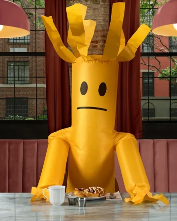 Inflatable tube man with sad face sits at marble table inside a restaurant with pastries, a mug, and condiments.